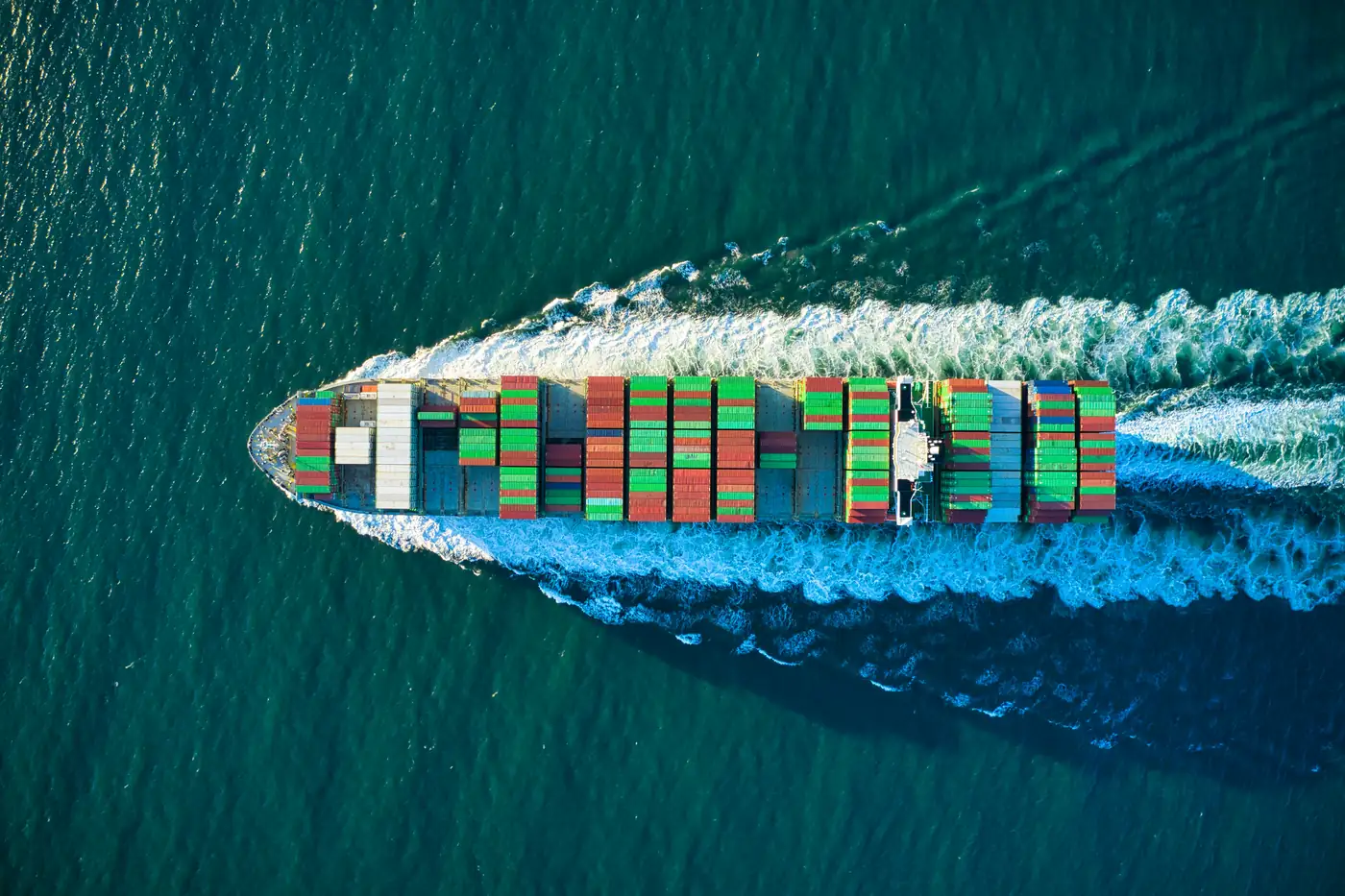 Et lastet containerskib set fra luften med et hvidt kølvand efter sig i grønt hav